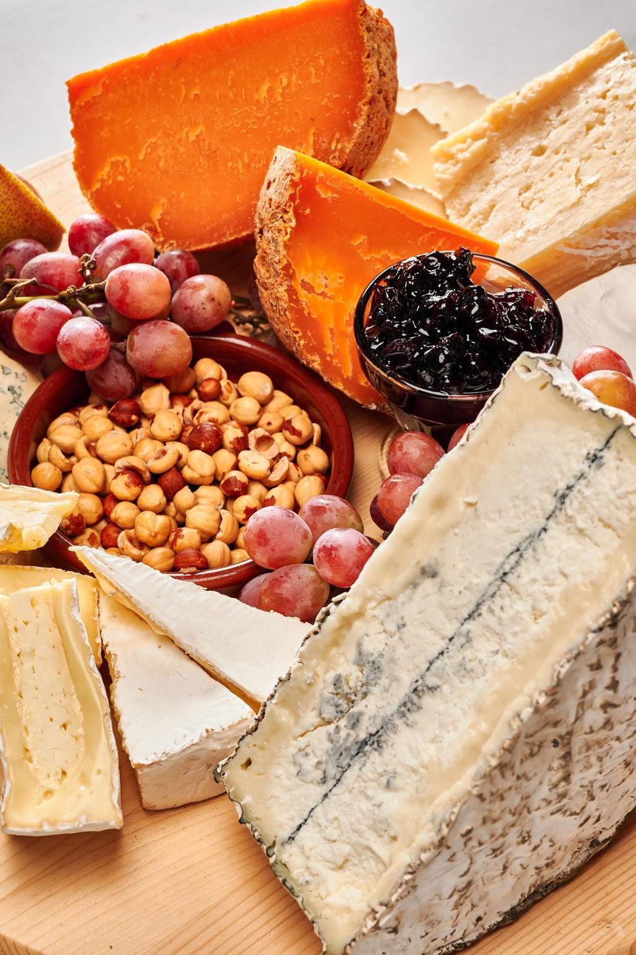 a cheese board on a round wooden platter set against a marble background. The board features a variety of cheeses, each with distinct colors and textures. There is an orange-hued aged cheese, a creamy brie or camembert, a wedge of blue cheese, and several other types, ranging from hard to soft in texture