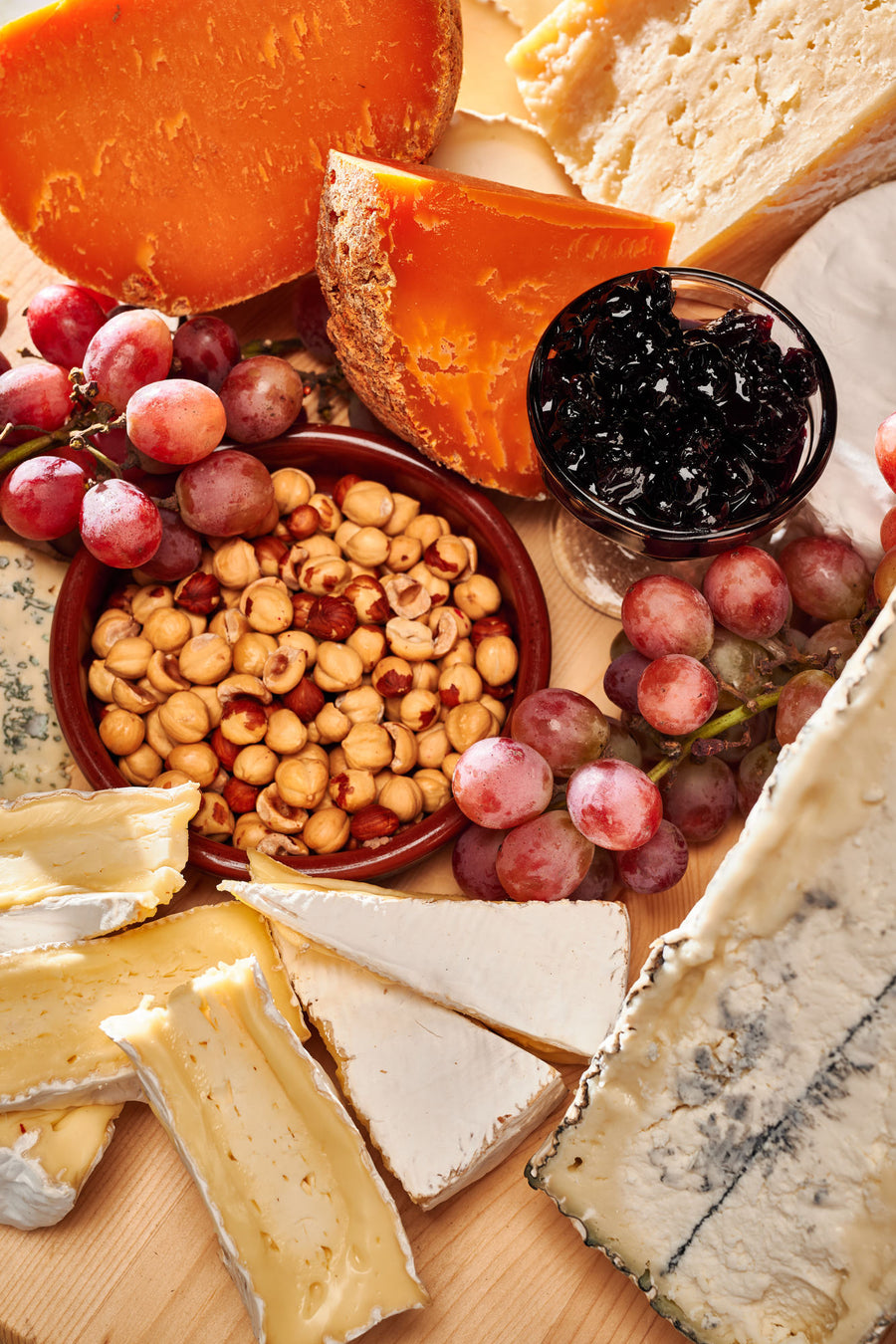a cheese board on a round wooden platter set against a marble background. The board features a variety of cheeses, each with distinct colors and textures. There is an orange-hued aged cheese, a creamy brie or camembert, a wedge of blue cheese, and several other types, ranging from hard to soft in texture