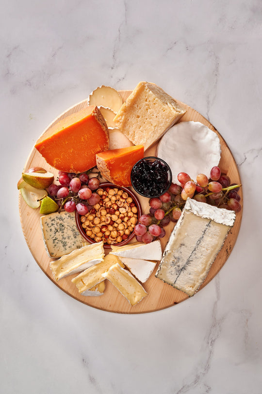 a cheese board on a round wooden platter set against a marble background. The board features a variety of cheeses, each with distinct colors and textures. There is an orange-hued aged cheese, a creamy brie or camembert, a wedge of blue cheese, and several other types, ranging from hard to soft in texture