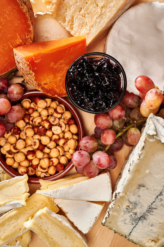 a cheese board on a round wooden platter set against a marble background. The board features a variety of cheeses, each with distinct colors and textures. There is an orange-hued aged cheese, a creamy brie or camembert, a wedge of blue cheese, and several other types, ranging from hard to soft in texture