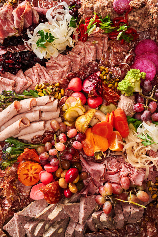 an olympia provions chefs choice board including but not limited to, ham, grapes, assortmens of salami, raisins, pistachios, potatos, carrots, olives, and pomegranates