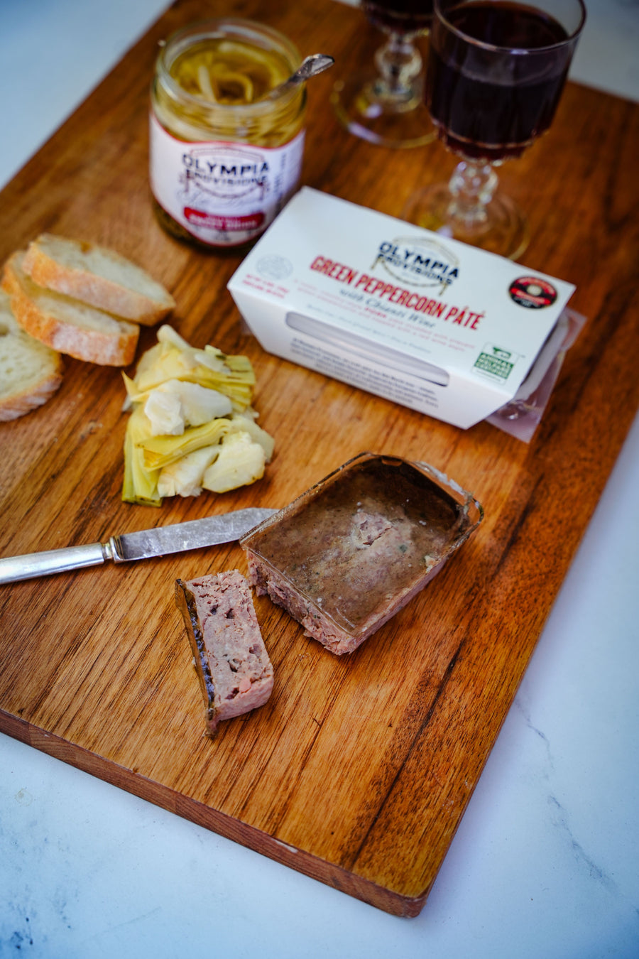 Green peppercorn pate, jar of pickles, and bread on a wooden cutting board with a marble background.