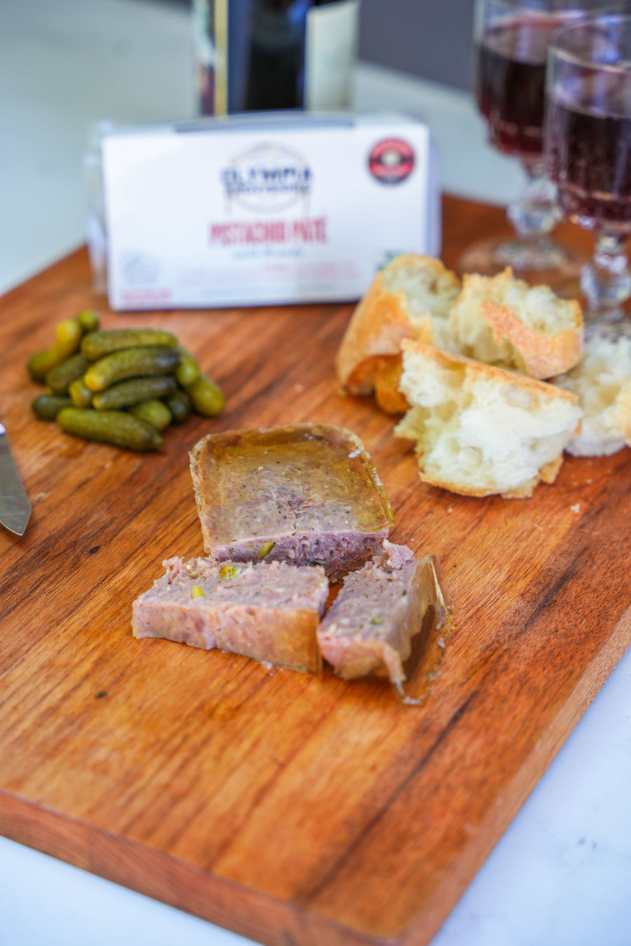 Wooden cutting board with sliced meat, bread, and pickles, with wine in the background.