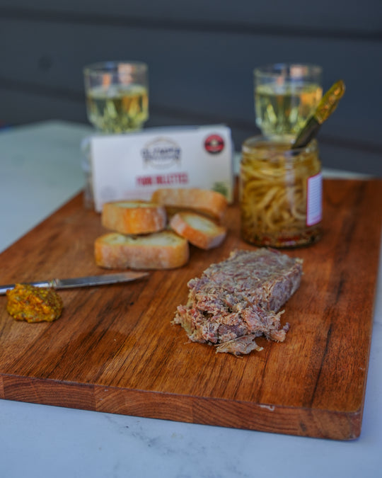 Wooden cutting board with sliced pork rillettes, bread, and a jar of pickles on a marble surface.