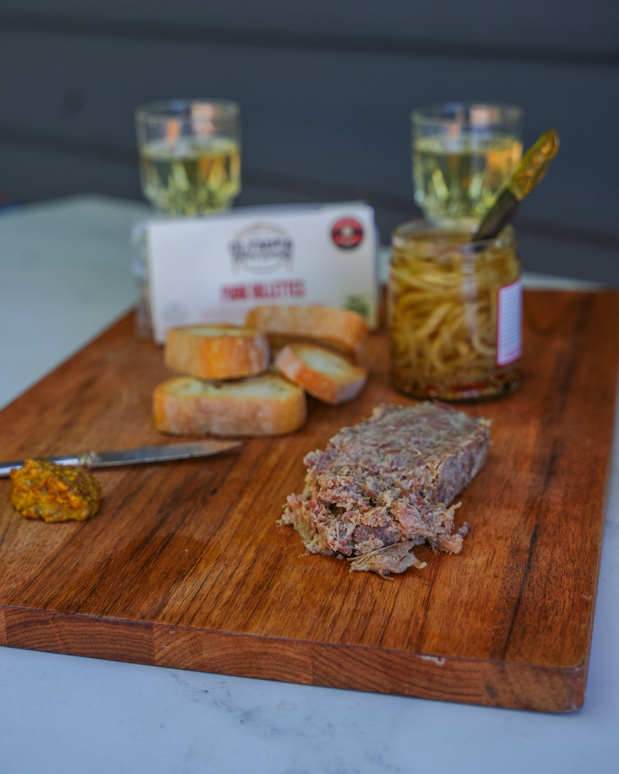 Wooden cutting board with sliced pork rillettes, bread, and a jar of pickles on a marble surface.