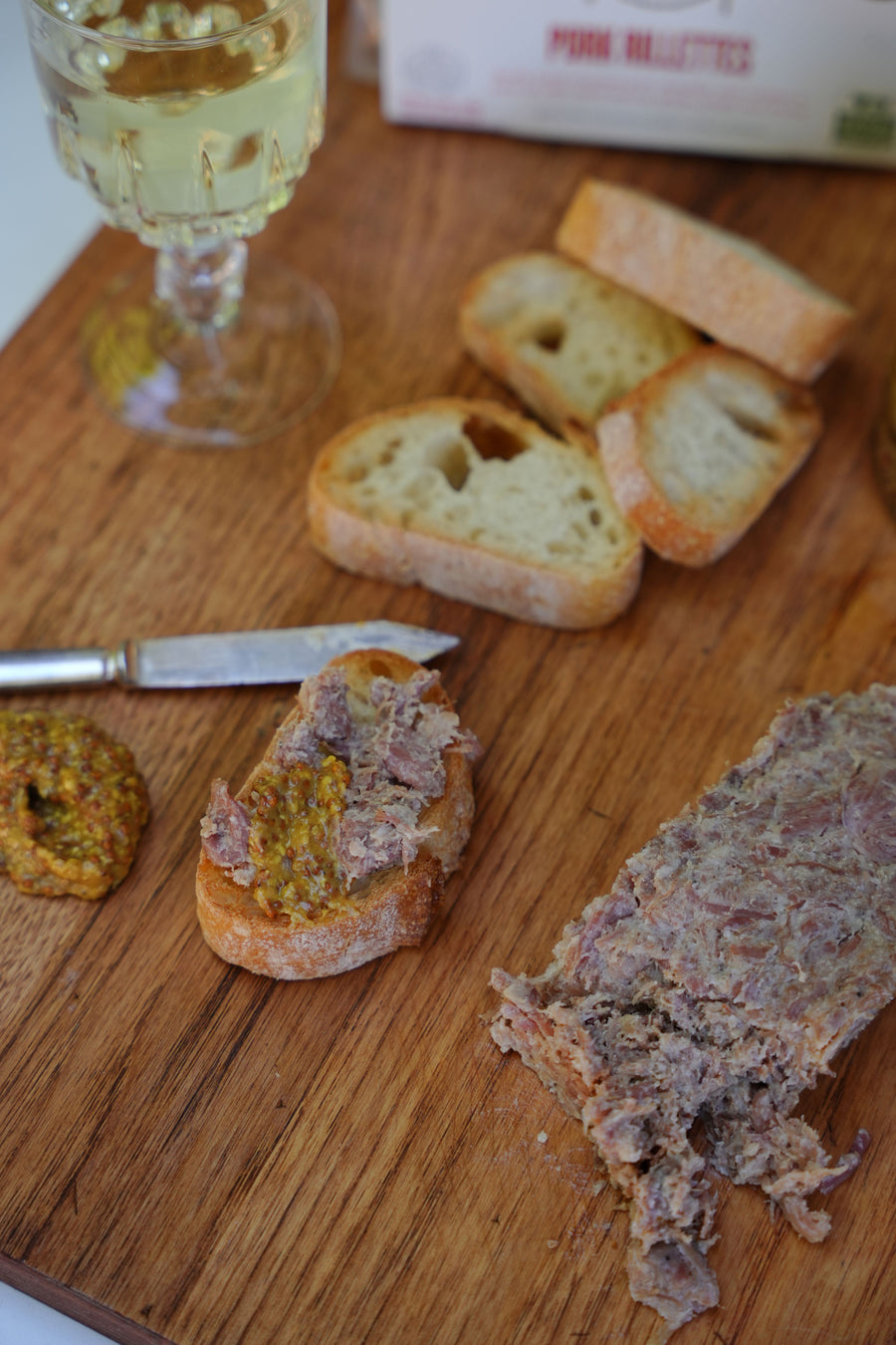 Wooden cutting board with bread slices, a loaf of pork rillettes, and a glass of white wine.