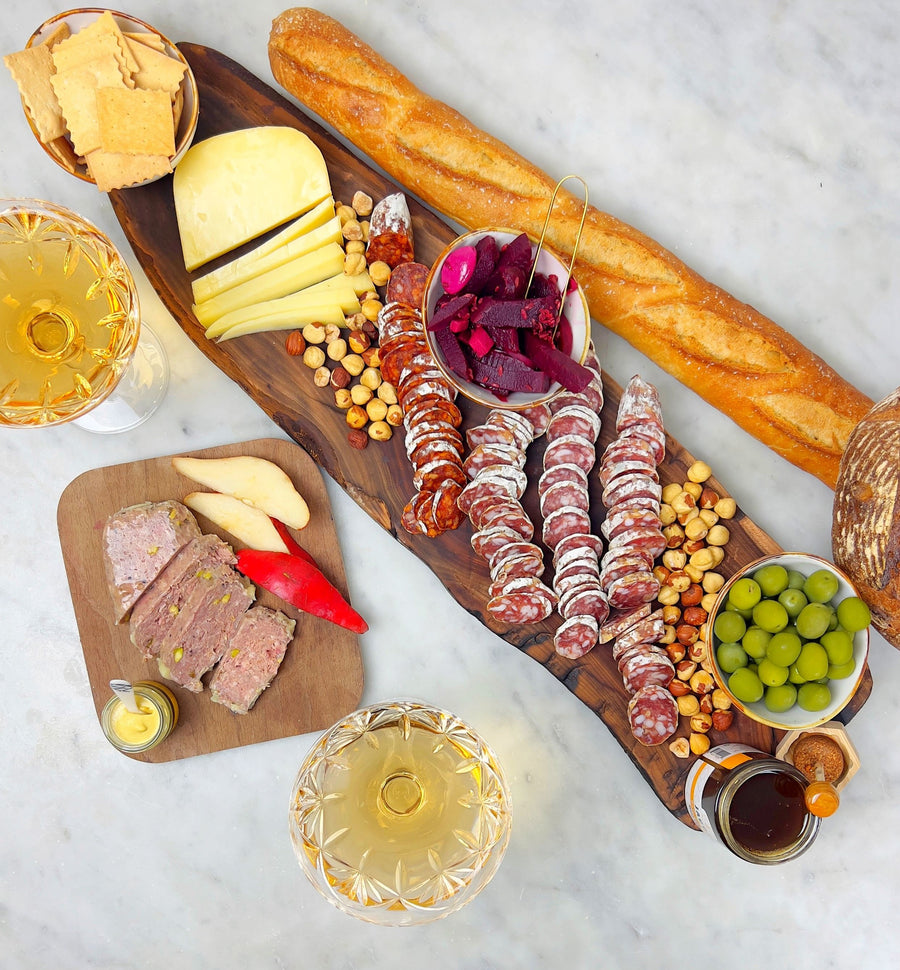 a charcuterie board of salami, hazlenuts, a baguette, a bowl of olives, a bowl of beets, and sliced cheese