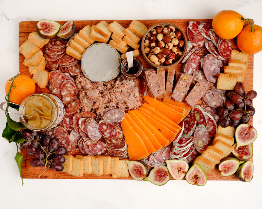 Wooden charcuterie board with various meats, cheeses, fruits, and nuts on a white background.