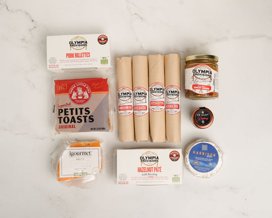 Collection of salami, petite toasts, pork rilletes, pate, cheese, pickled onions, and chees on a white surface.