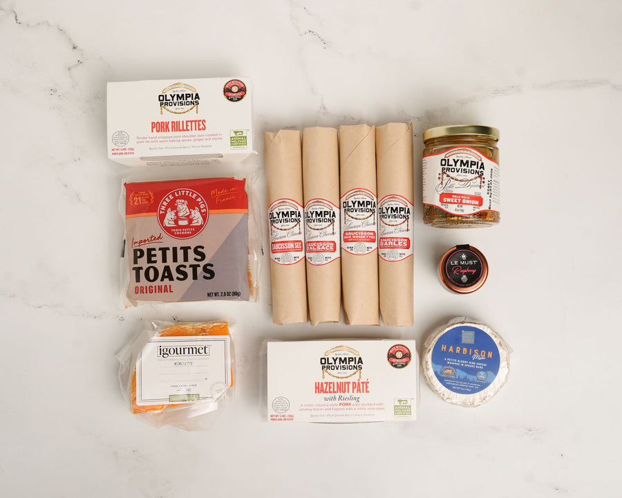 Collection of salami, petite toasts, pork rilletes, pate, cheese, pickled onions, and chees on a white surface.