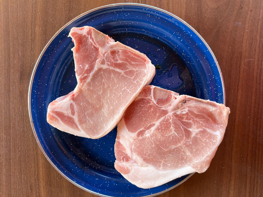 Two raw pork chops resting on a dark blue speckled ceramic plate, set on a brown wooden table.