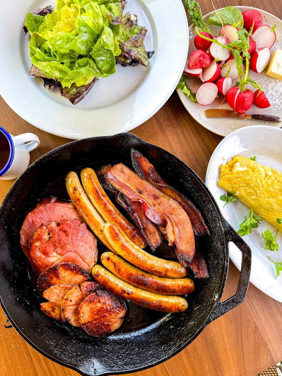 A skillet filled with grilled sausages, bacon, and ham served on a wooden table. Surrounding the skillet are plates with a fresh green salad, radishes, and an omelette, along with a coffee cup