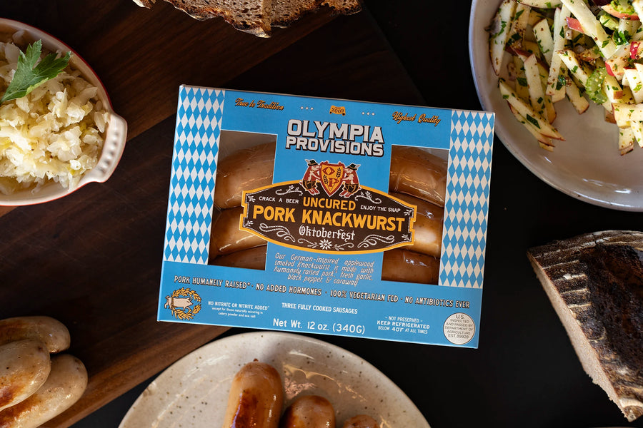 One pack of Olympia Provisions Pork Knackwurst sausages surrounded by plates of food on a wooden table.