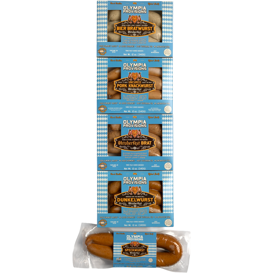 Five packages of Olympia Provisions Oktoberfest sausages stacked vertically in blue and white packaging. from top to bottom: Bier Bratwurst, Pork Knackwurst, Oktoberfest Brat, Dunkelwurst, and Speckwurst.