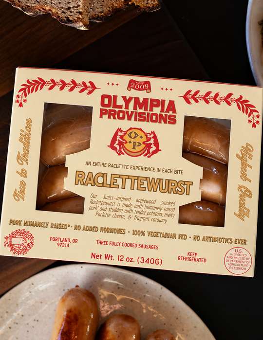 Three links of Olympia Provisions Raclettewurst sausage in cream packaging on a wooden table surrounded by plates of food.